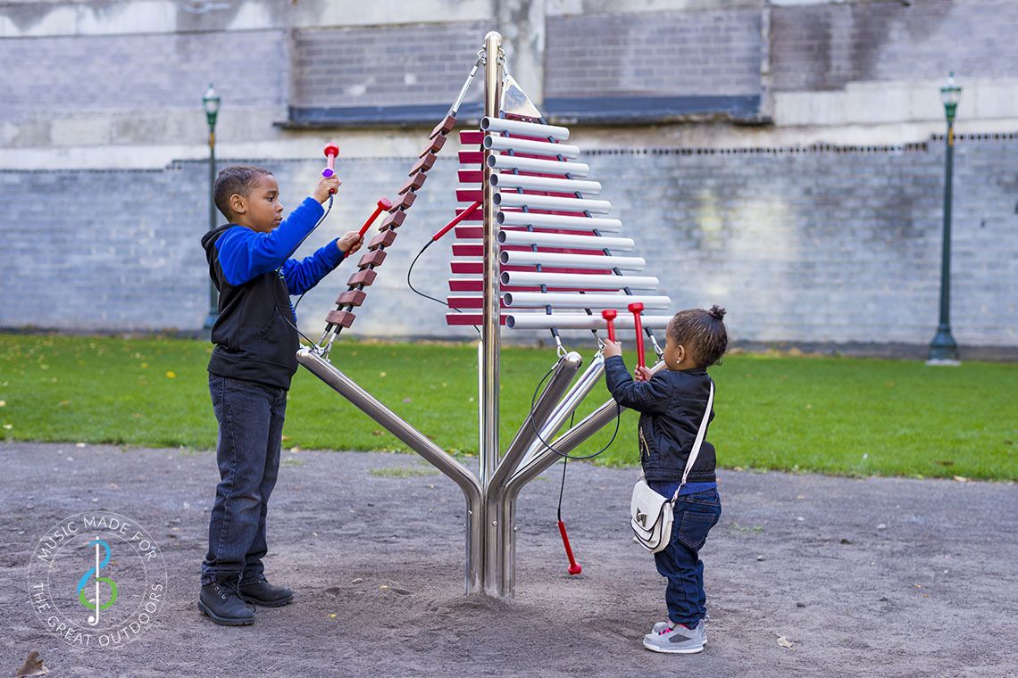 large outdoor musical instrument with one post and three vertical xylophones being played by one boy and one girl