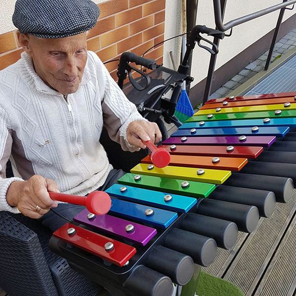 Elderley man sat playing a large colourful xylophone in a courtyard garden