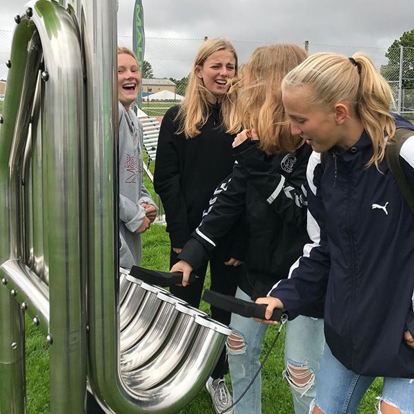Group of teenage girls in a playground laughing and hitting a large silver outdoor musical instrument with five tall pipes 