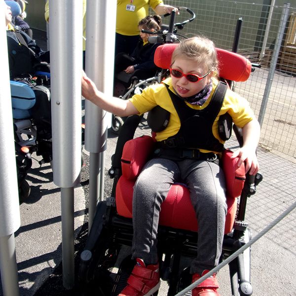 girl in wheelchair reaching out and touching large tubular bell in school playground