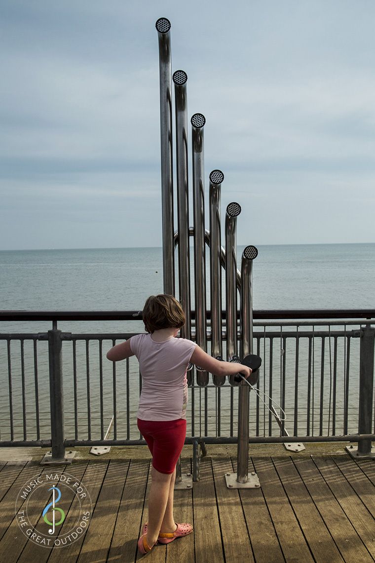 back of girl hitting large silver outdoor aerophones or slap tubes on the end of a pier looking out to sea
