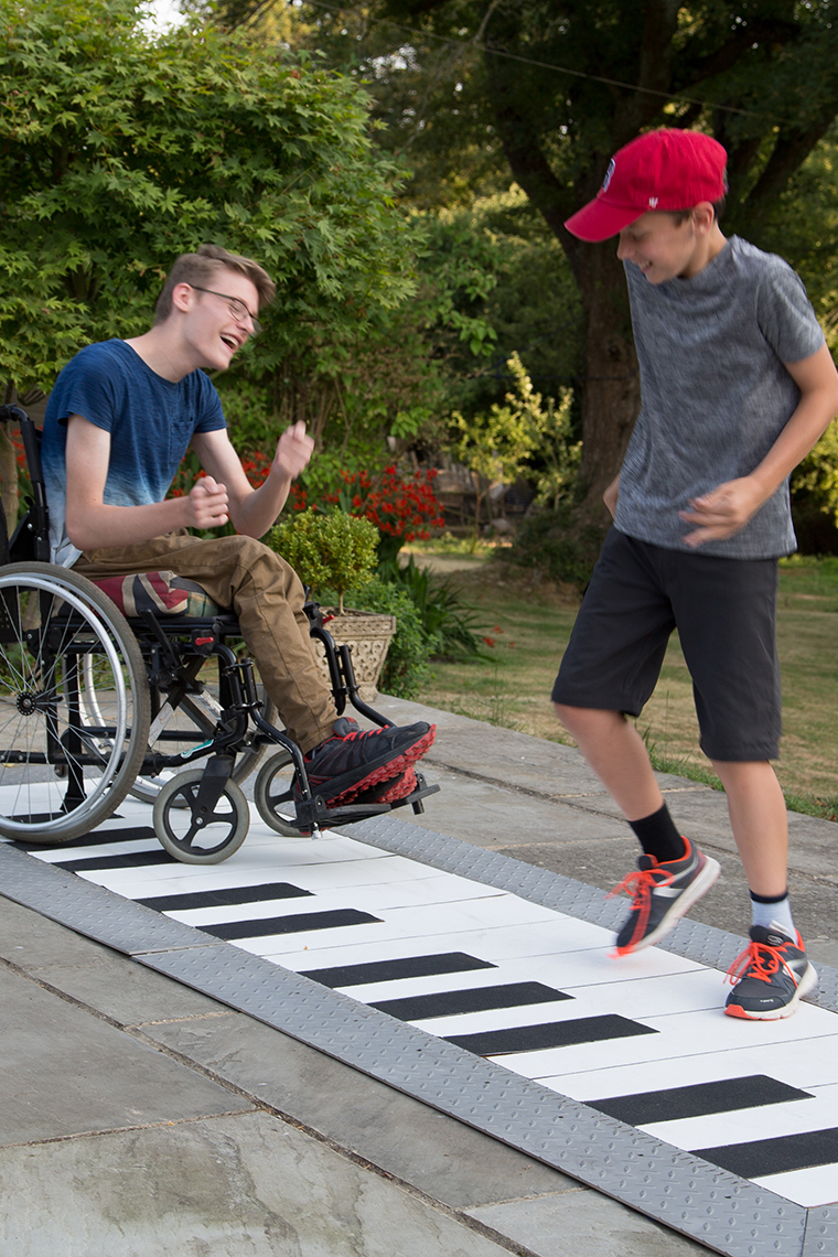 two boys, one in a wheelchair playing on an outdoor floor piano in a park