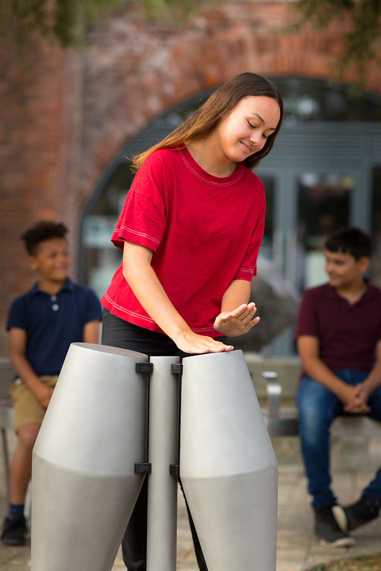 a young black girl in a red t shirt is playing a pair of steel tumbadora drums in a street watched by her two brothers