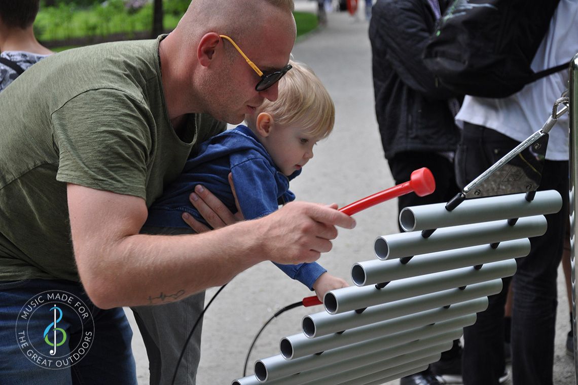 father holding a small child up and playing a large outdoor xylophone together in the park