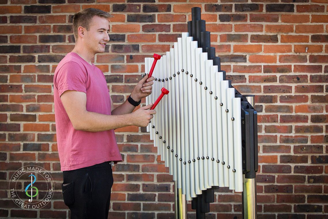 young man playing large outdoor musical instrument with upright tubular notes mounted on black resonators