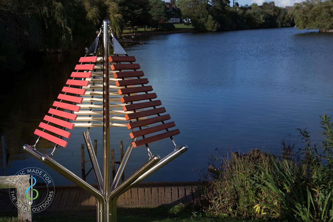 large outdoor musical instrument with one stainless steel post in ground with three vertical xylophones attached by lakeside