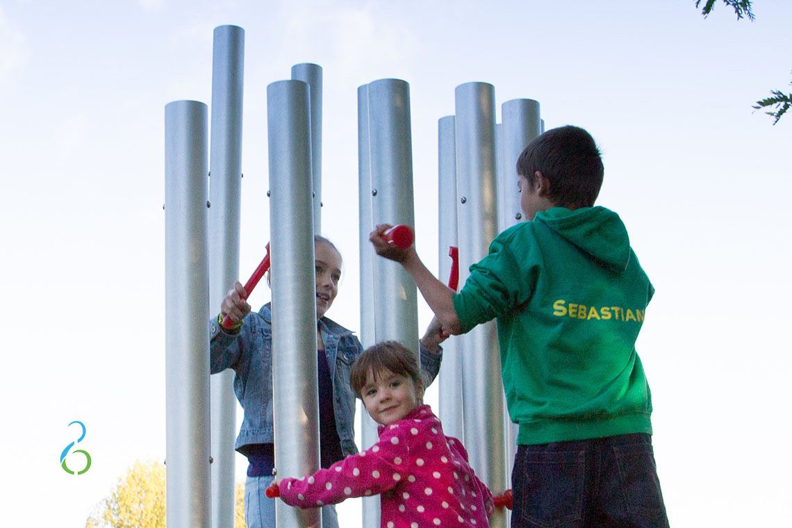 Three children hitting large silver coloured outdoor chimes with red beaters