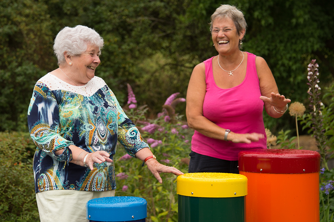 two older ladies playing a colourful set of 5 outdoor samba drums in a park